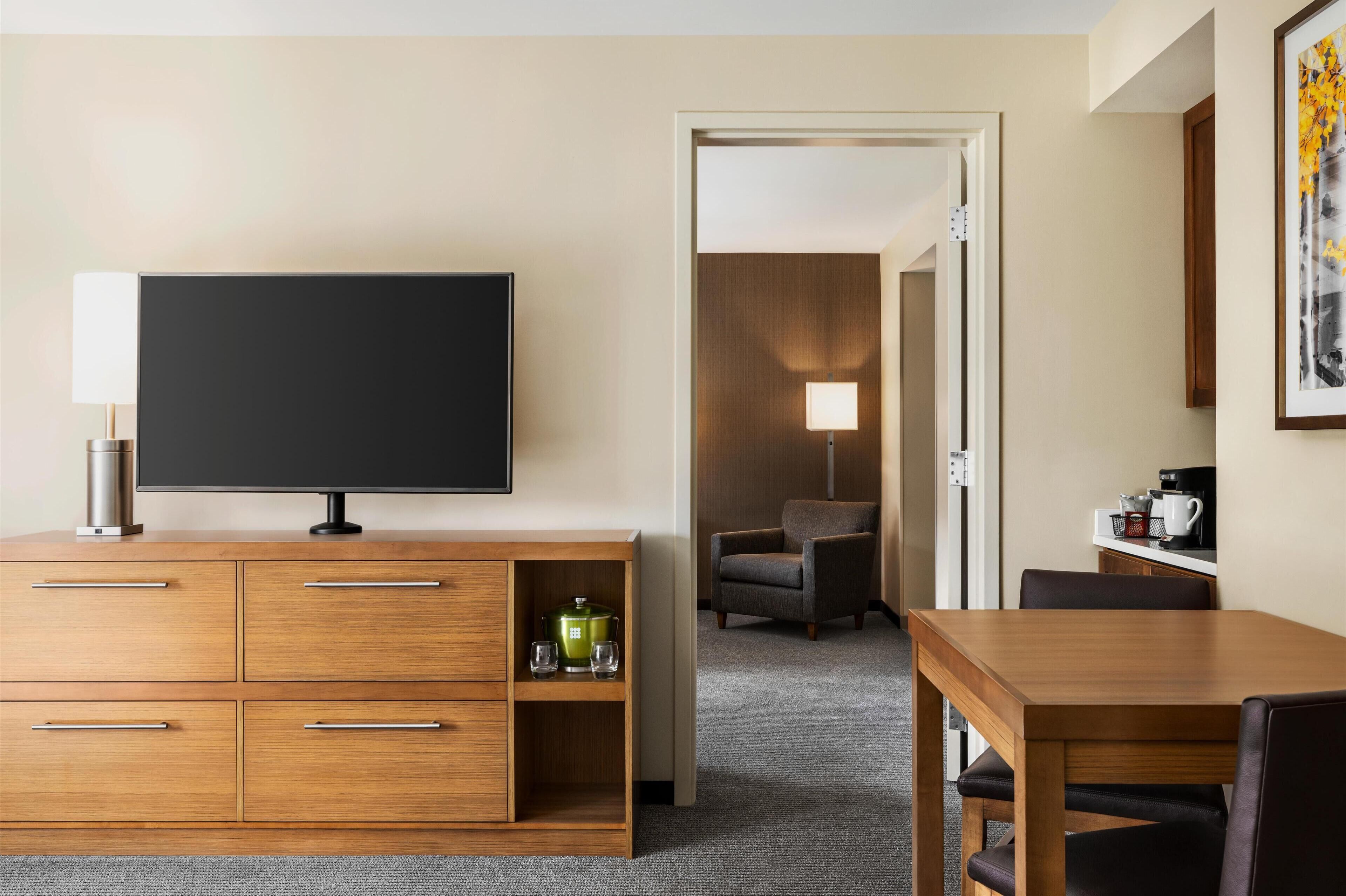 A hotel room with a flat-screen TV on a wooden cabinet, a dining table with chairs, a lamp, and a coffee maker.