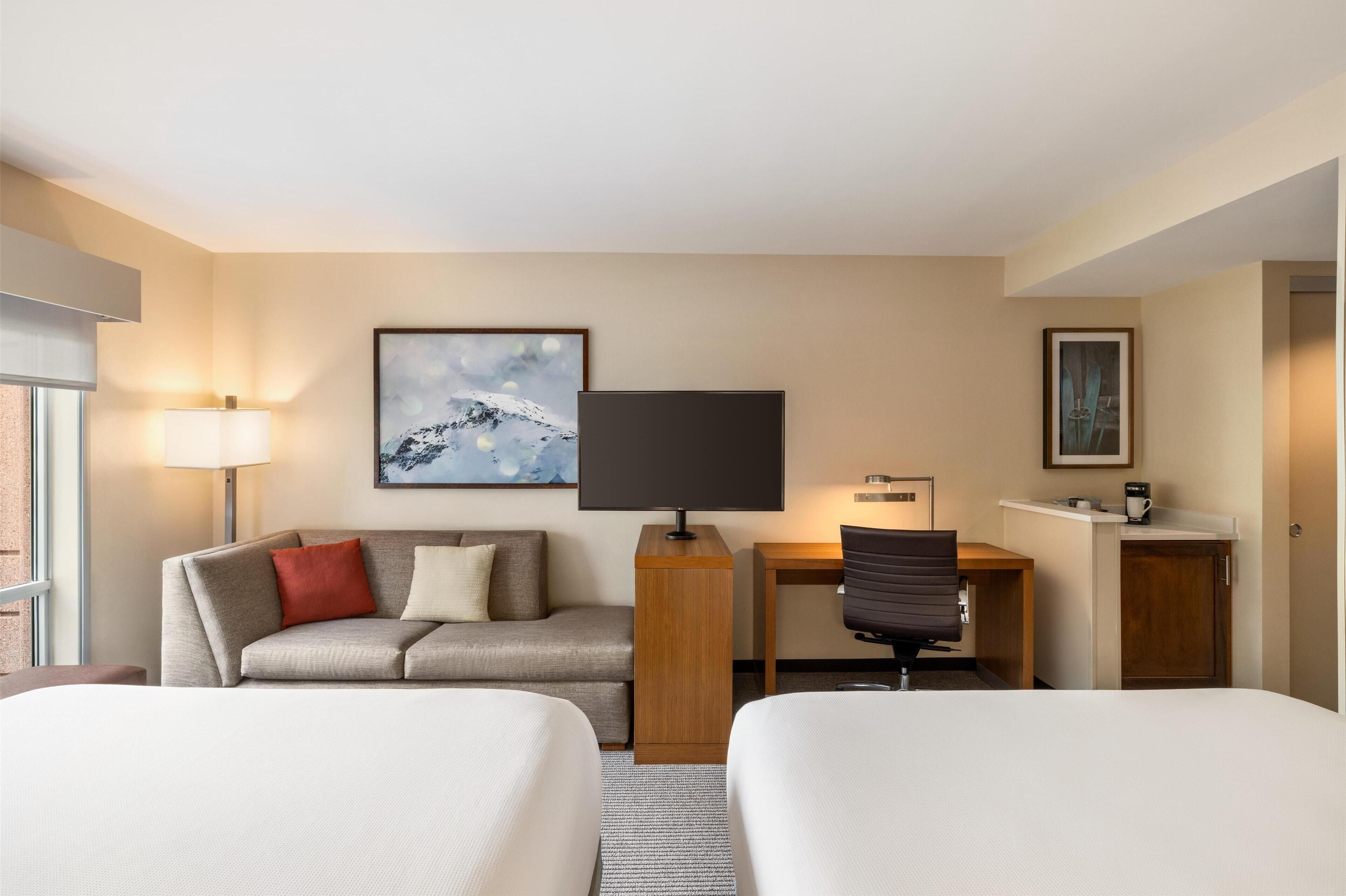 Une chambre d’hôtel avec deux lits, un bureau, un canapé, une lampe et des œuvres d’art aux murs.
