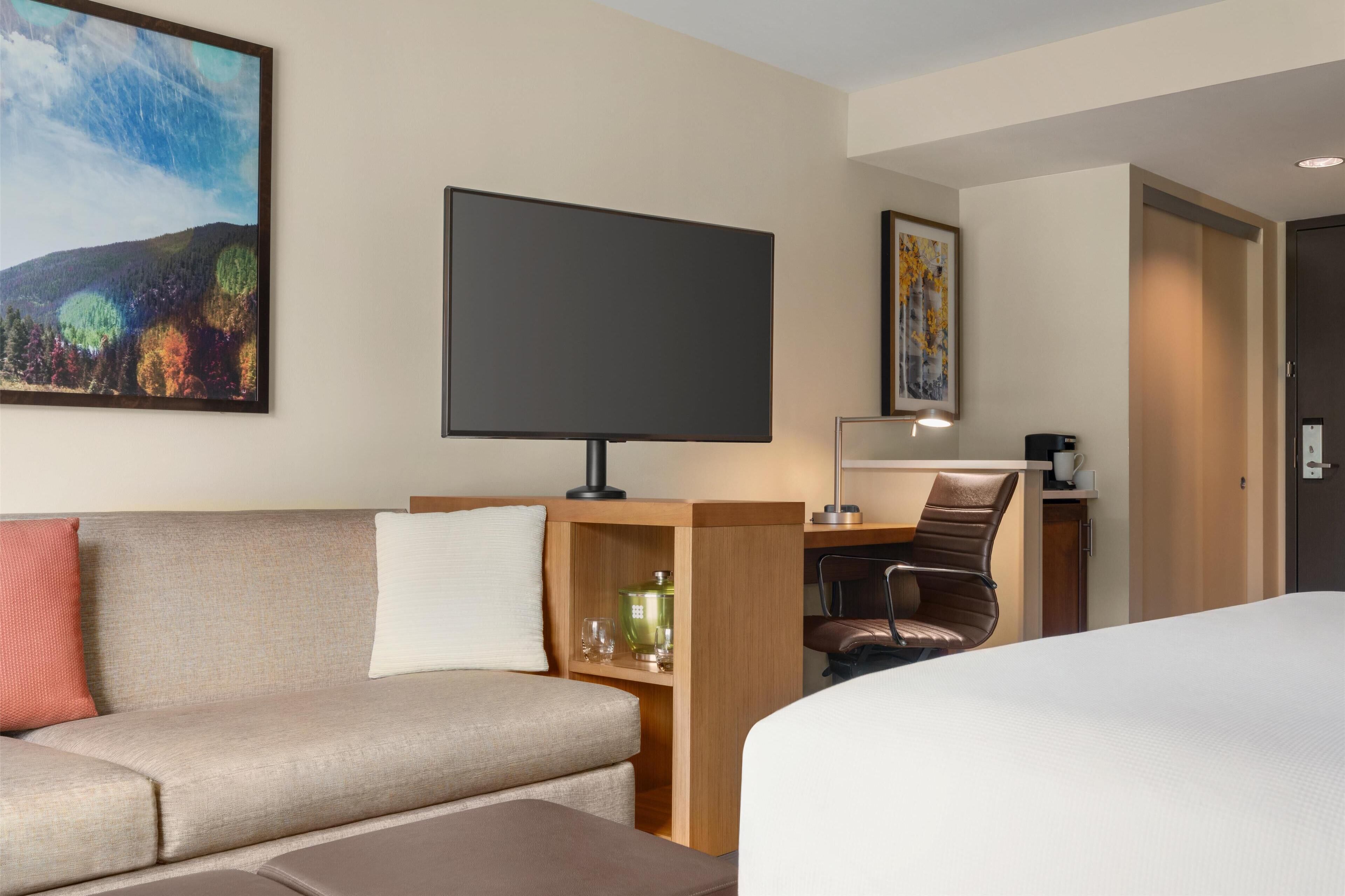 A hotel room with a bed, a sofa, a desk with a computer, a chair, a TV, and a painting on the wall.