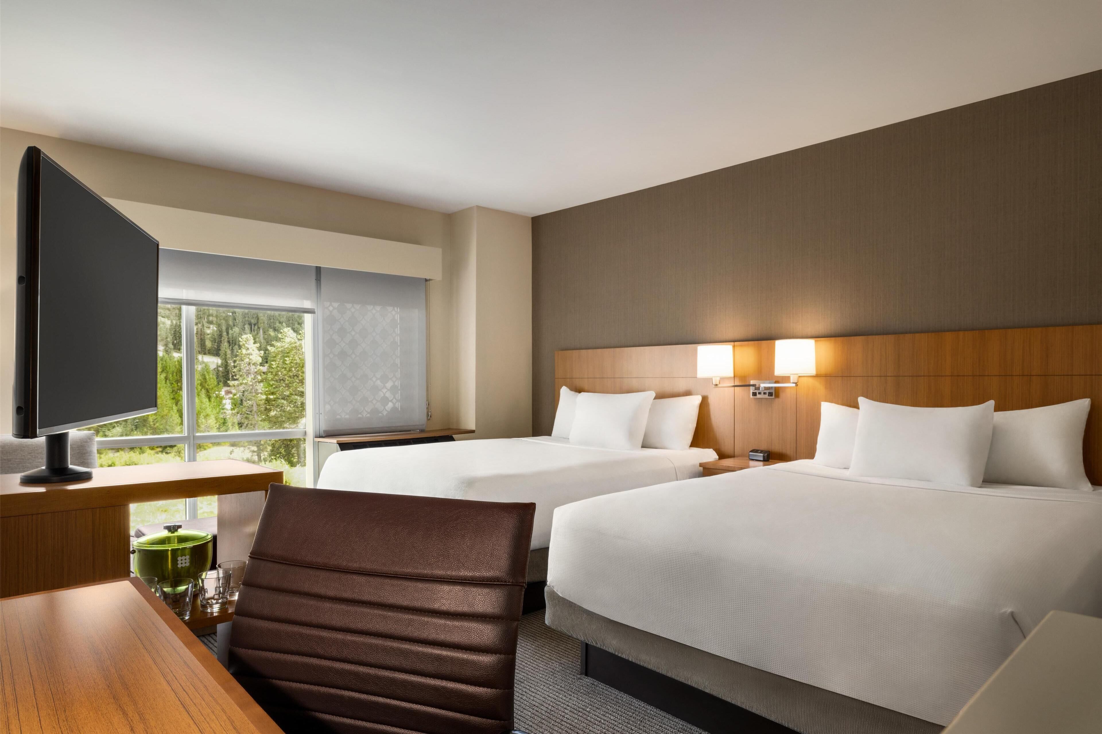 A hotel room with two beds, a desk with a television, and a window with a view of trees.