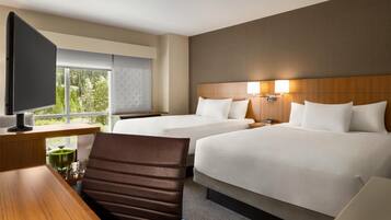 A hotel room with two beds, a desk with a television, and a window with a view of trees.