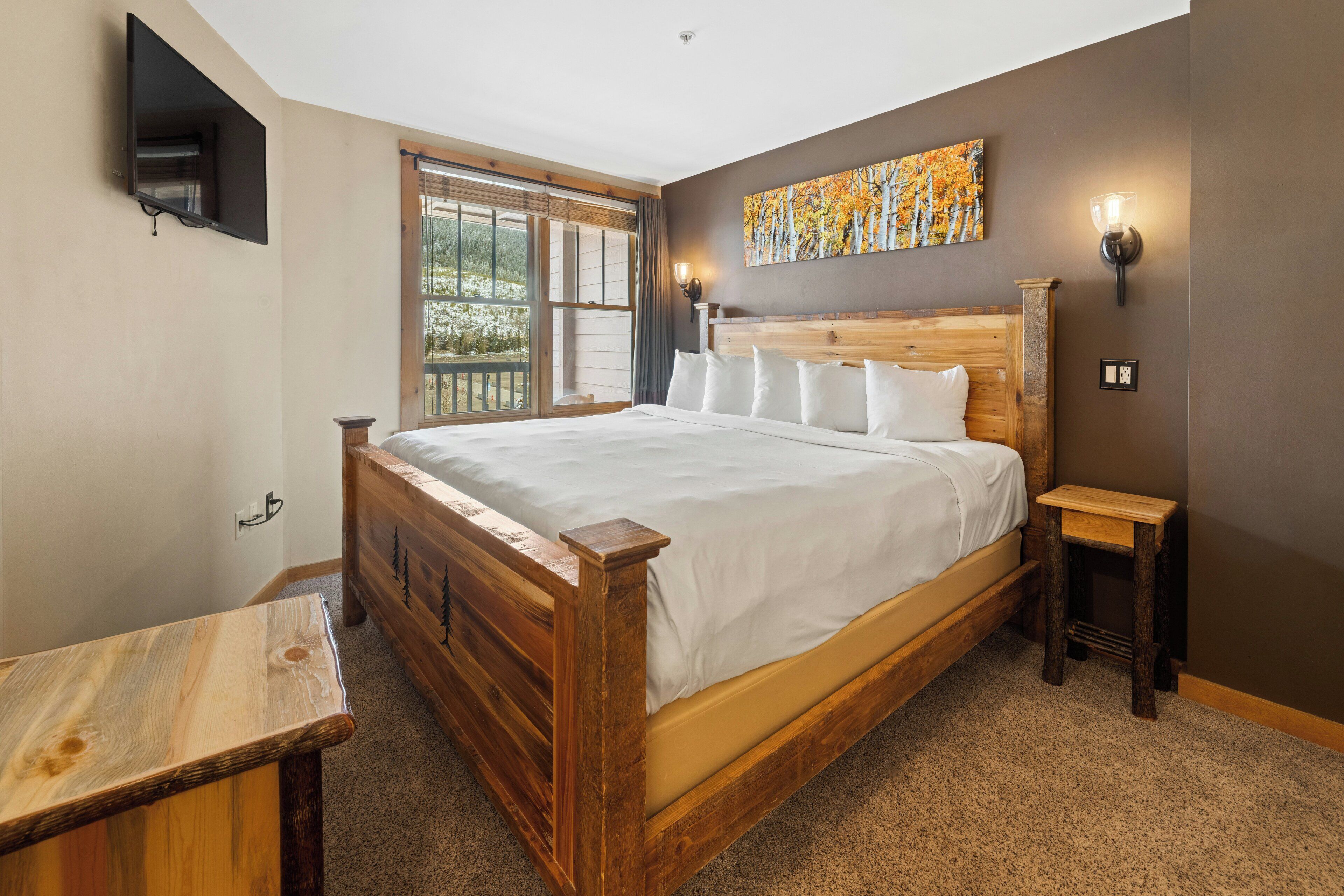 A bedroom with a wooden bed, a small wooden table, a TV mounted on the wall, and a window with a view of greenery.