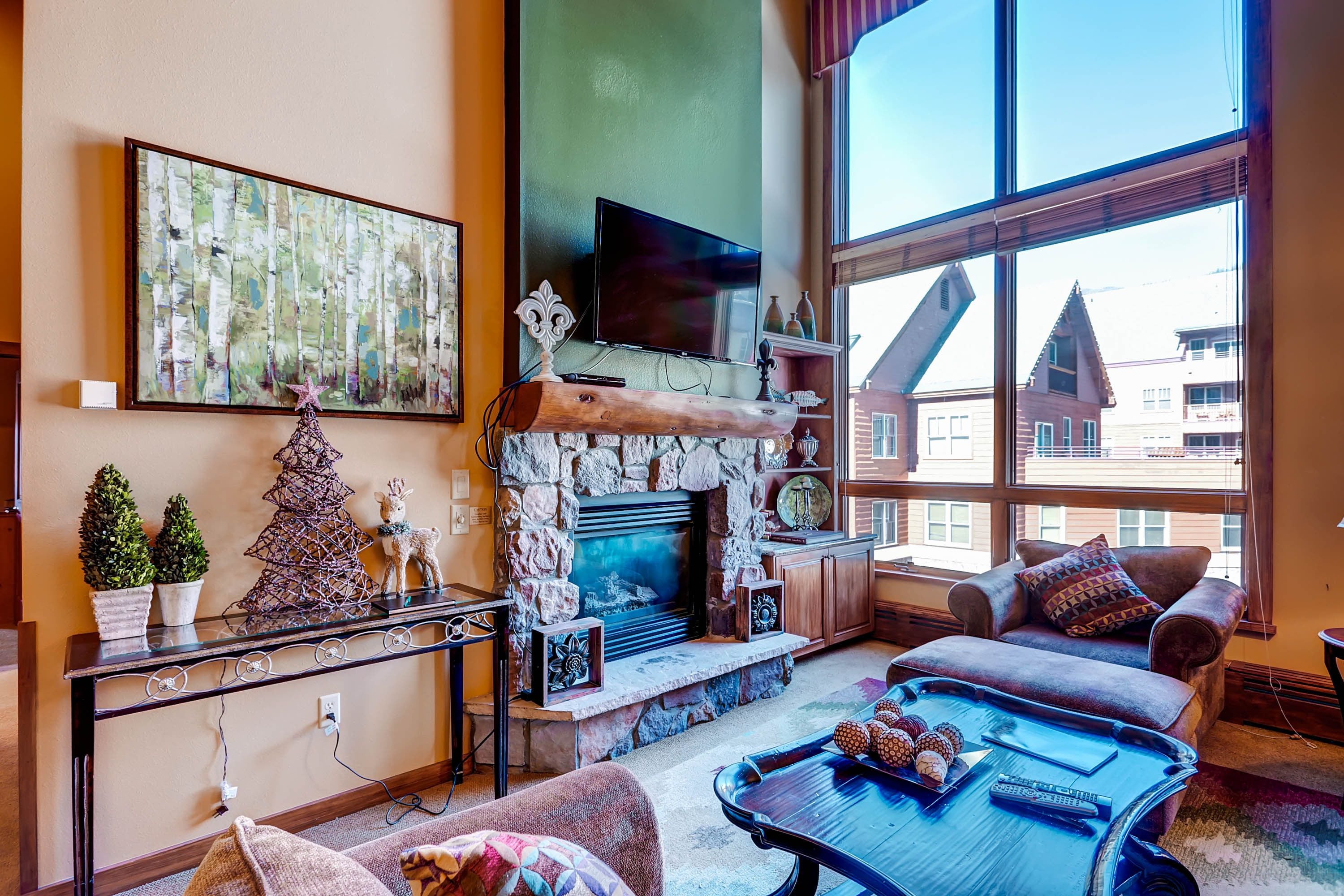 A cozy living room with a stone fireplace, a large window with a view of buildings, a comfortable sofa, and a coffee table with decorative items.
