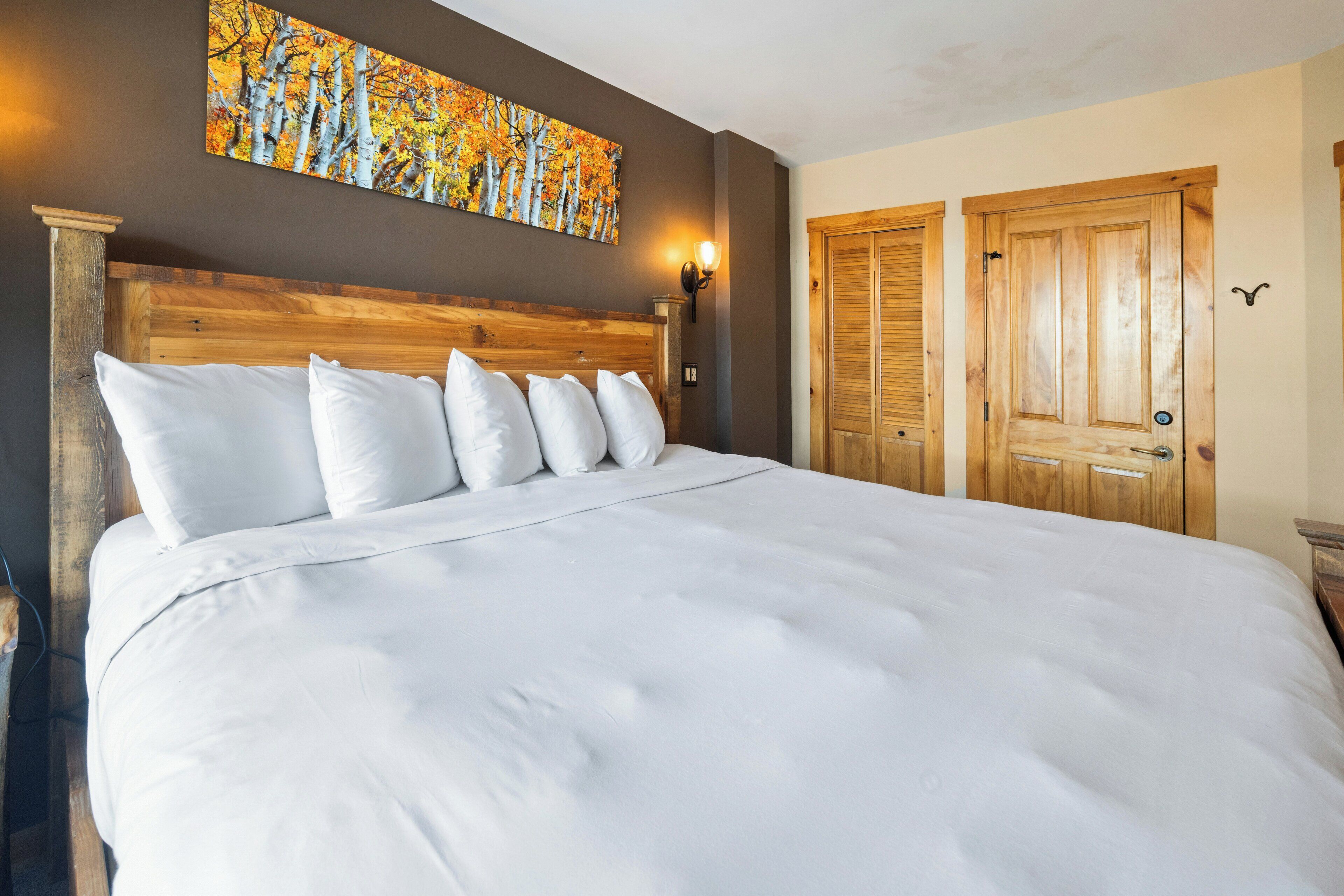 A large bed with white linens, a wooden headboard, and a wall-mounted painting of autumn trees.