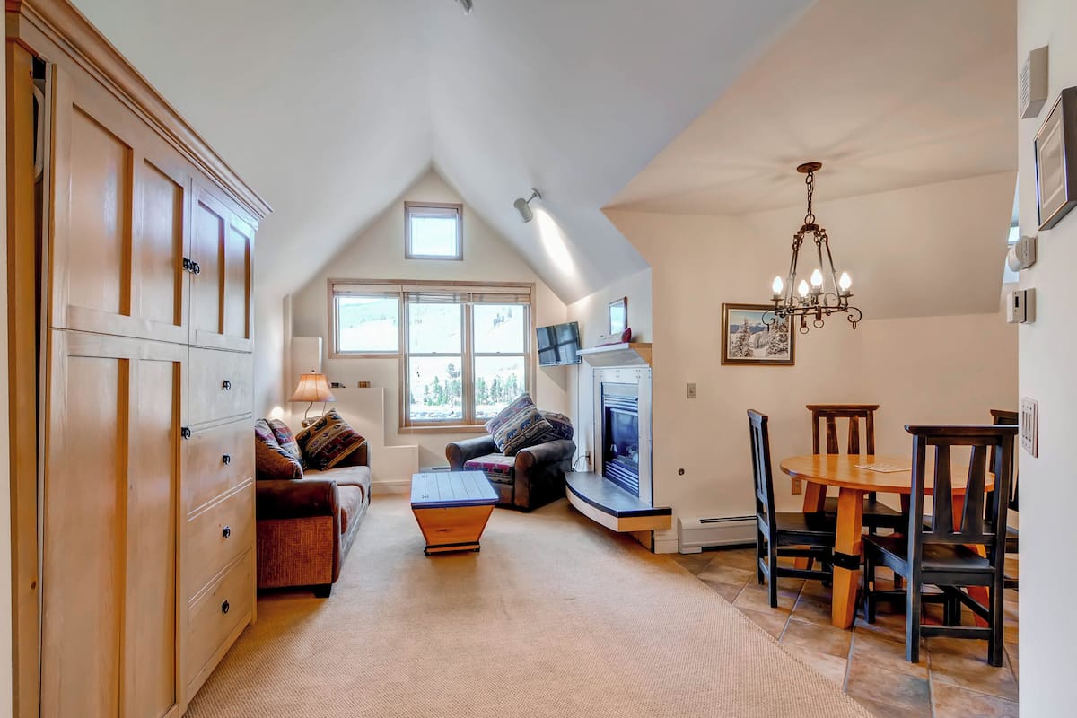 A cozy living room with a fireplace, a dining area with wooden chairs, and a large window providing natural light.