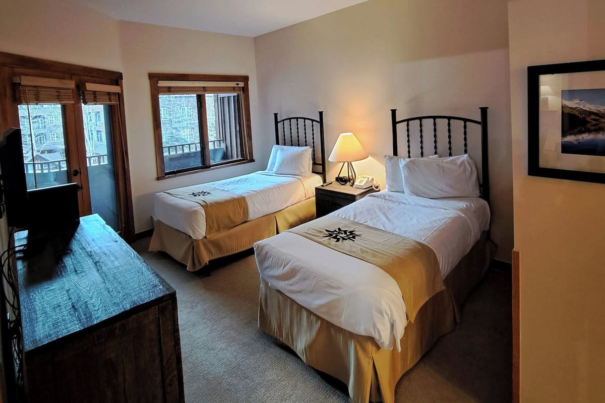 A hotel room with two beds, a desk, a lamp, and a television.