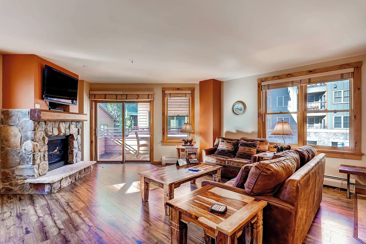 A spacious living room with a stone fireplace, wooden furniture, and large windows offering a view of the outside.