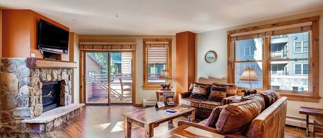 A spacious living room with a stone fireplace, wooden furniture, and large windows offering a view of the outside.