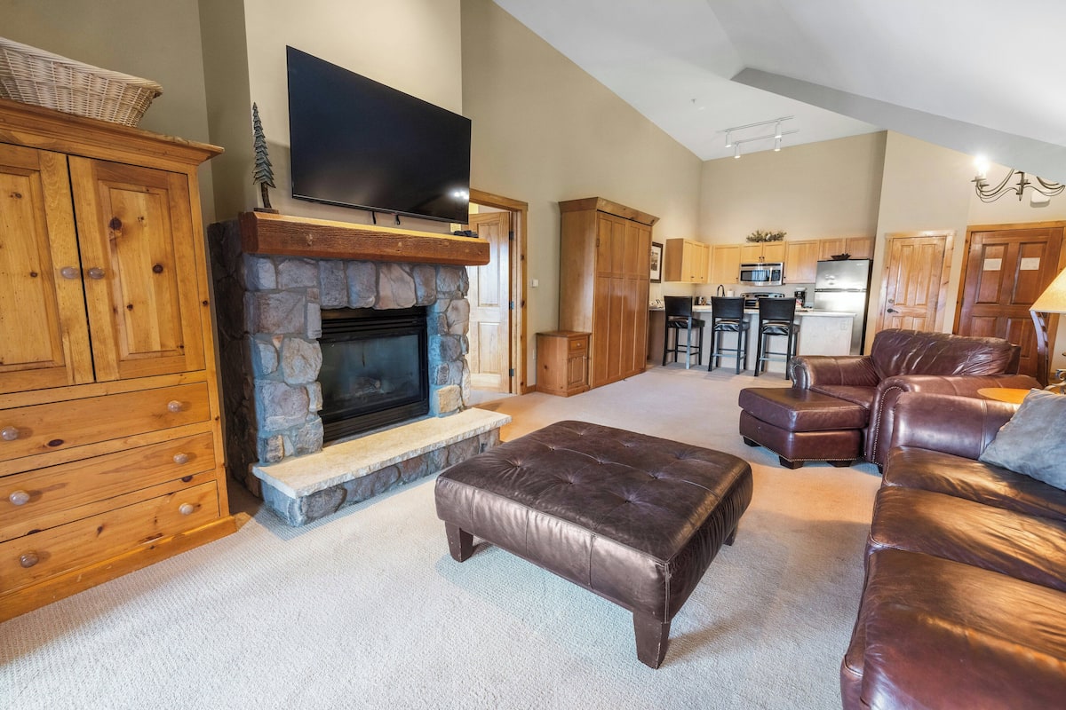A living room with a stone fireplace, leather furniture, a flat-screen TV, and a kitchen in the background.