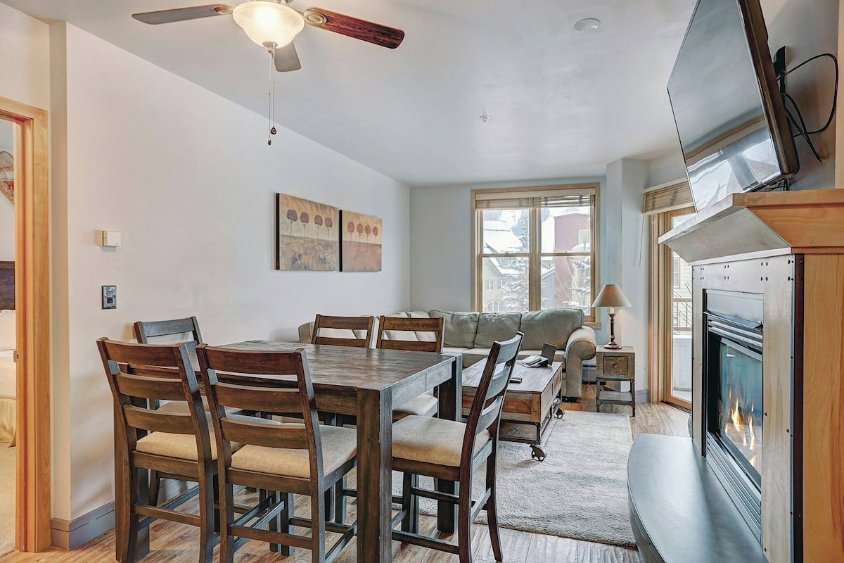 A dining area with a wooden table and chairs, a sofa, a fireplace, and a ceiling fan.