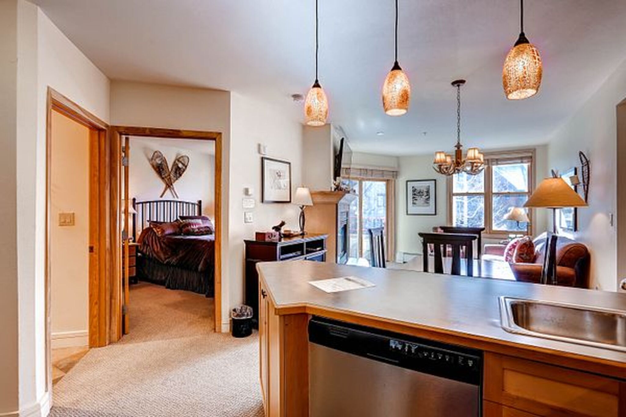 A modern kitchen with a dishwasher, a wooden island, pendant lights, and a view of a bedroom with a bed and a fireplace.