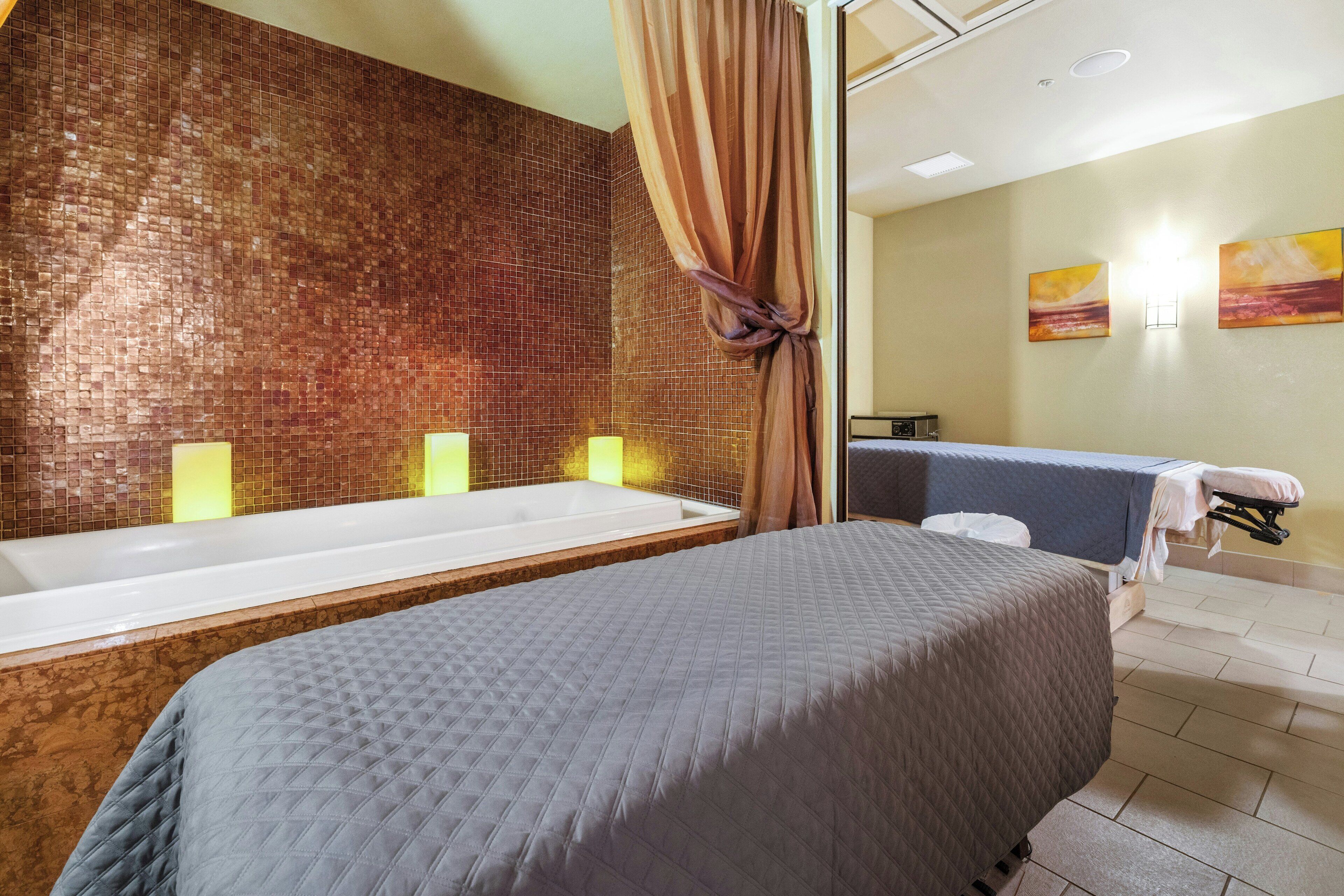 Une chambre d’hôtel dotée d’une grande baignoire, d’un lit recouvert d’une courtepointe grise et d’un mur orné de motifs en mosaïque.