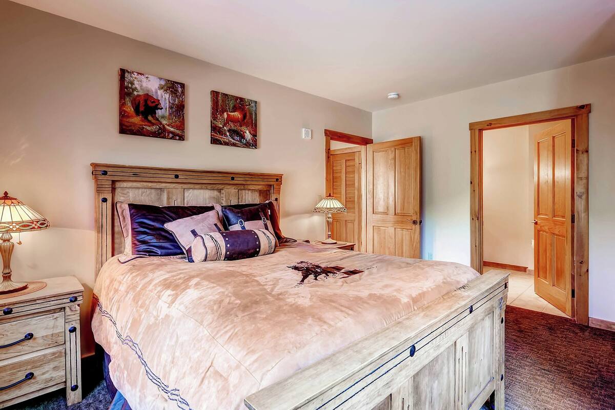 A bedroom with a large bed, wooden headboard, two paintings on the wall, and a wooden door.