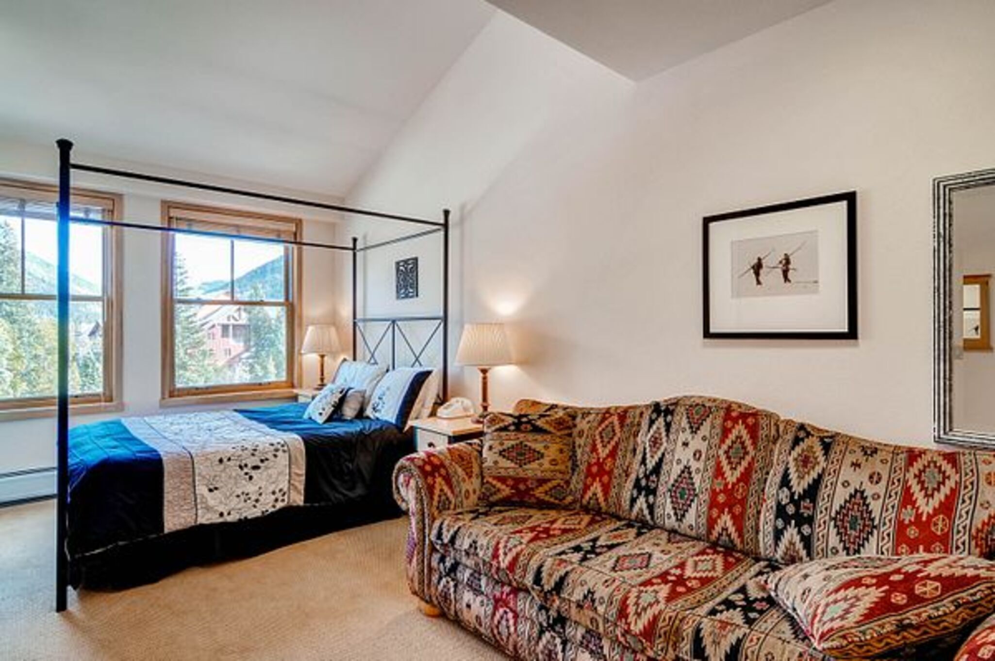 A bedroom with a four-poster bed, a patterned sofa, a framed picture on the wall, and a window with a view of trees.