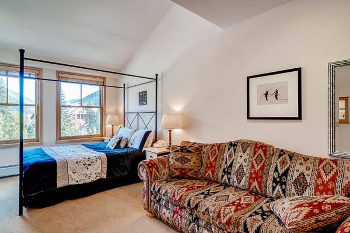 Une chambre à coucher avec un lit à baldaquin, un canapé à motifs, un tableau encadré au mur et une fenêtre donnant sur des arbres.
