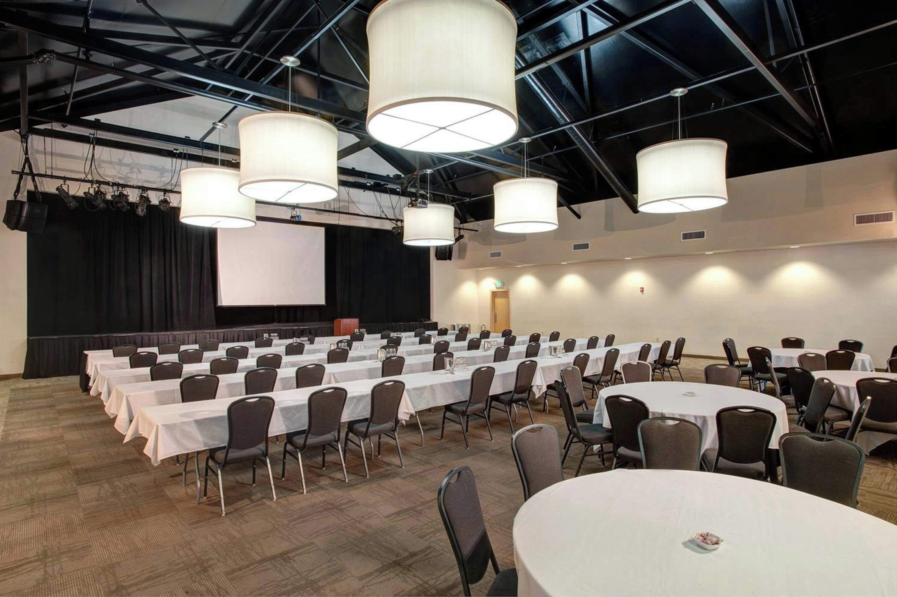 A conference room with multiple tables and chairs, a stage with a screen, and large hanging lights.