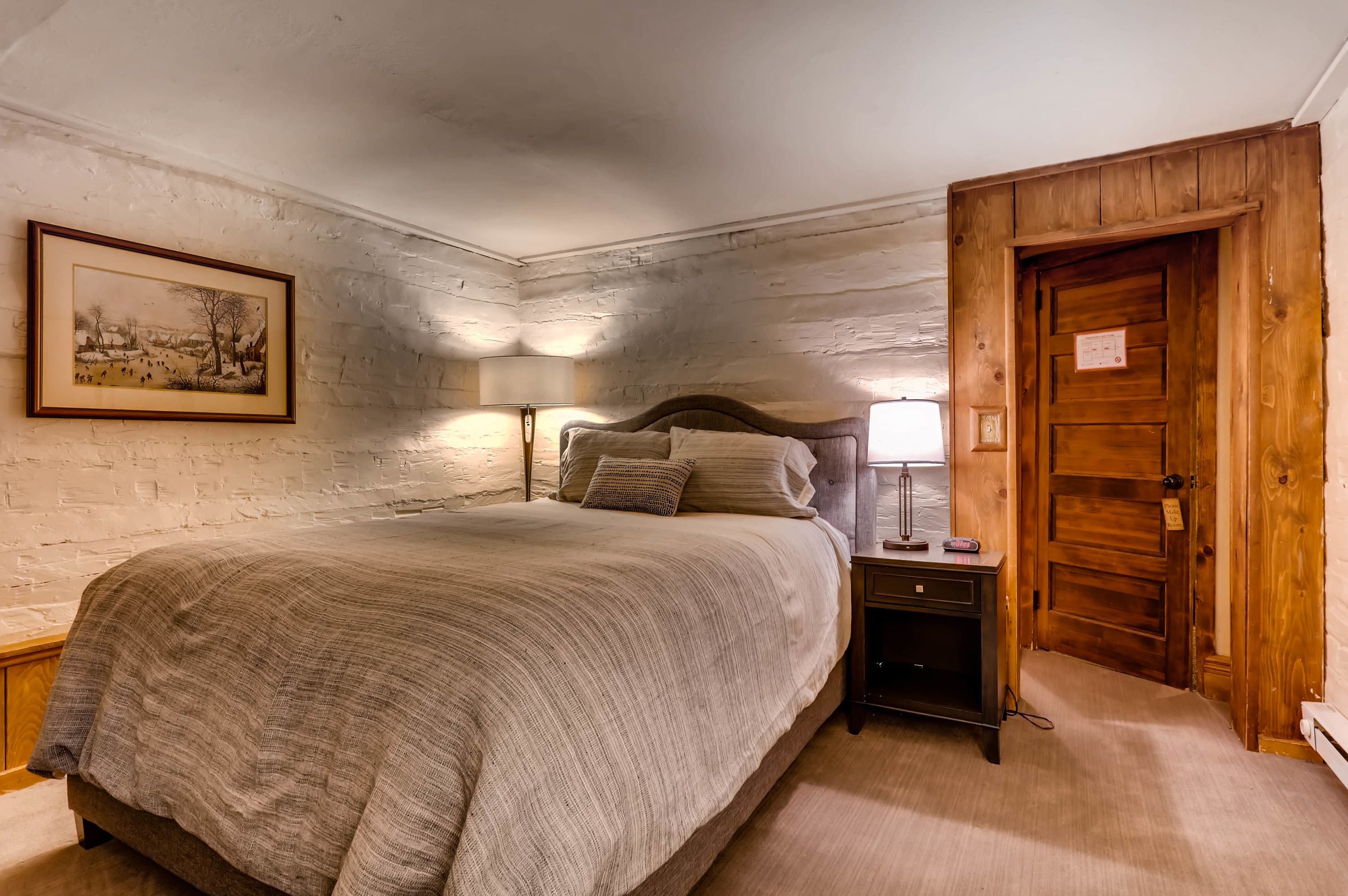 A bedroom with a large bed, a wooden door, a nightstand with a lamp, and a framed picture on the wall.