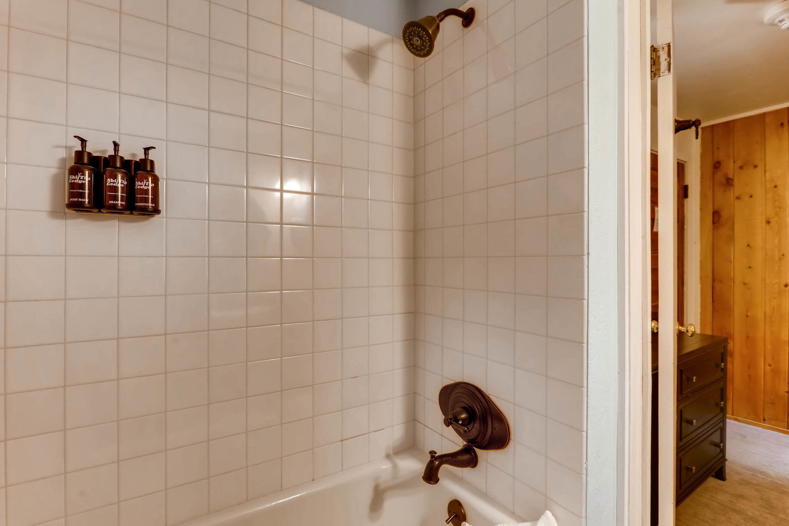 A bathroom with white tiled walls, a bathtub, a showerhead, and three bottles of shampoo on the wall.