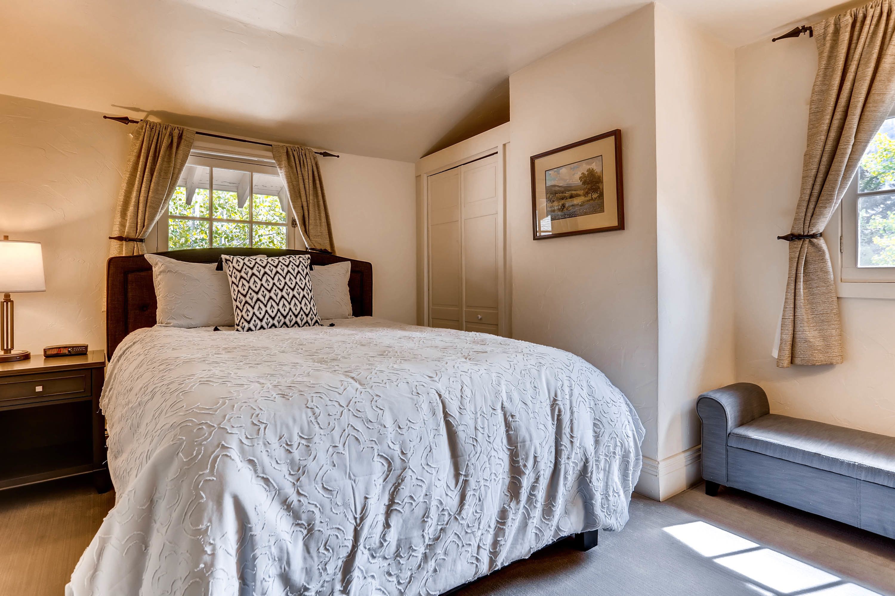 A bedroom with a large bed, a bedside table with a lamp, a bench, and a window with curtains.