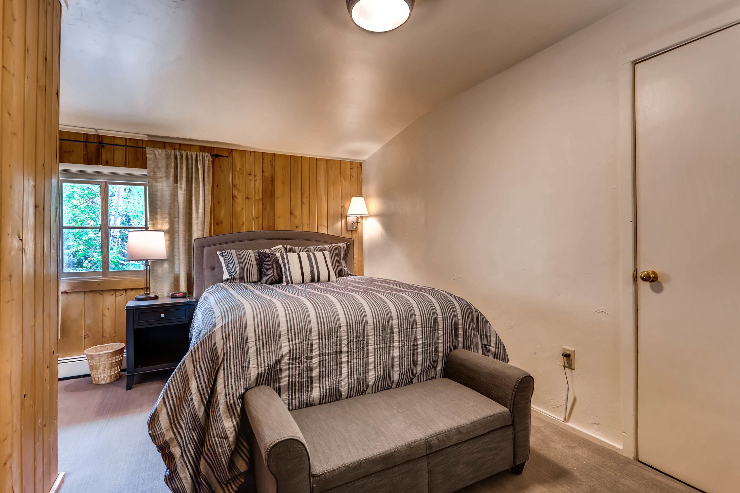 A bedroom with a bed, bedside lamp, wooden wall paneling, a window with curtains, and a door.