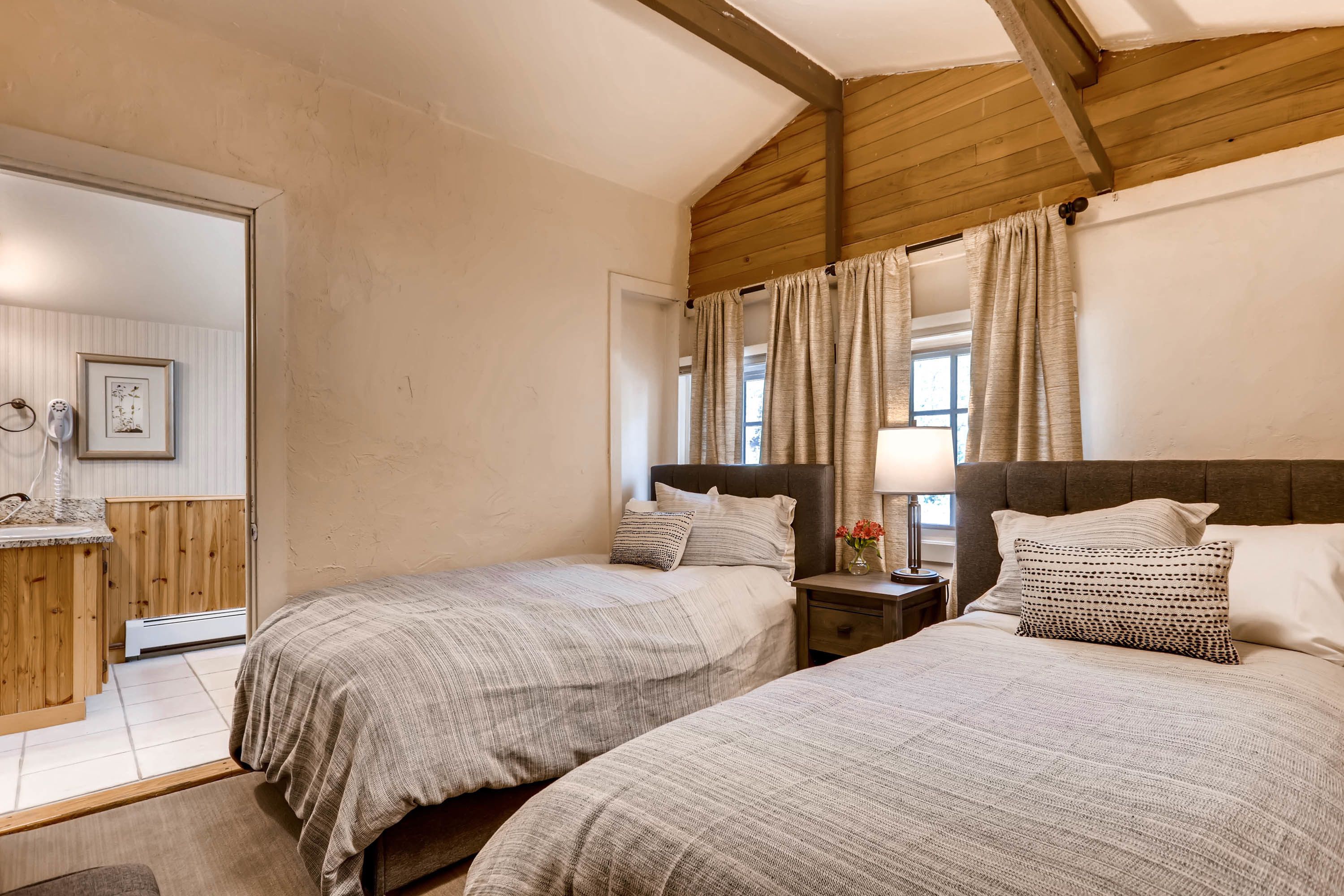 A bedroom with two beds, wooden beams on the ceiling, a nightstand with a lamp, and a window with curtains.