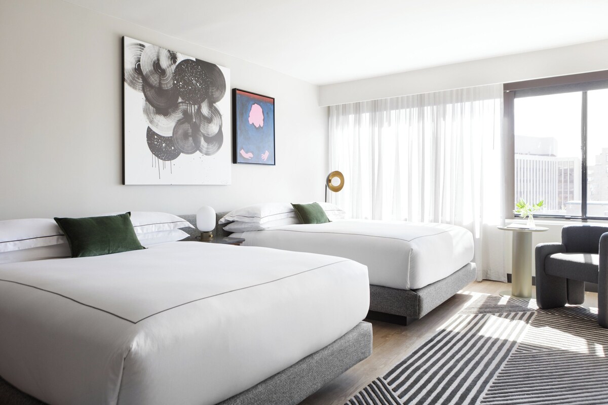 A modern hotel room with two beds, a small round table, a chair, and large windows with sheer curtains.