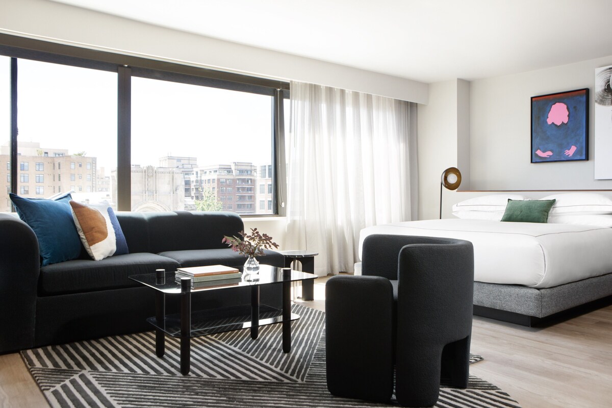 Un hotel moderno con un sofá negro, una cama blanca, una mesa de centro de vidrio y vistas a la ciudad a través de amplias ventanas.