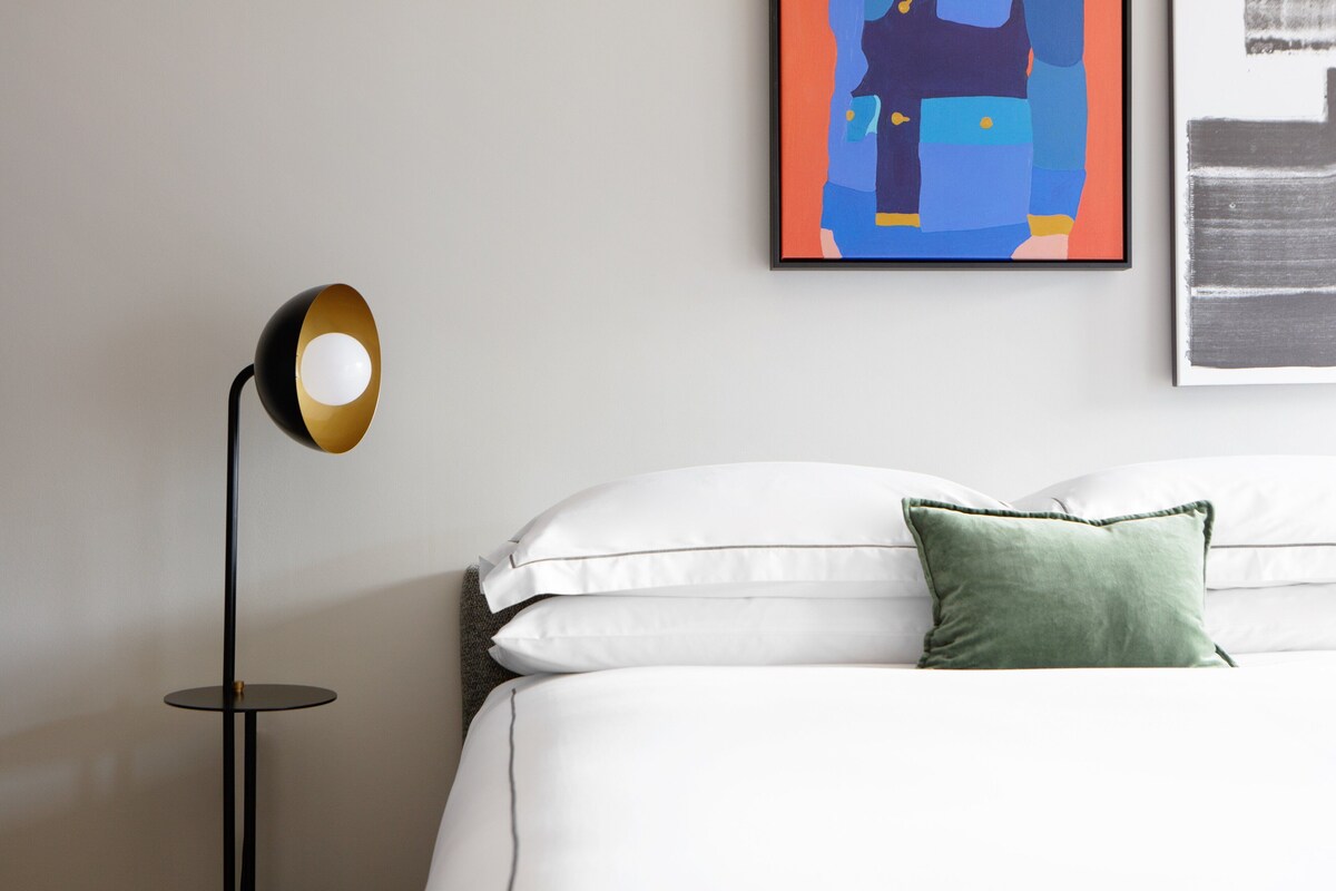 Una cama bien hecha con ropa blanca y una almohada verde, una lámpara de pie negra con base dorada, y un cuadro abstracto colorido en la pared.
