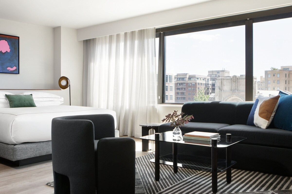 Una habitación de hotel moderna con una cama, un sofá negro, una mesa de centro de vidrio y vistas de la ciudad a través de amplias ventanas.
