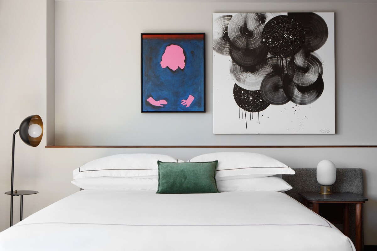 Un dormitorio moderno con cama, almohada verde, mesita de noche con lámpara y dos obras de arte en la pared.