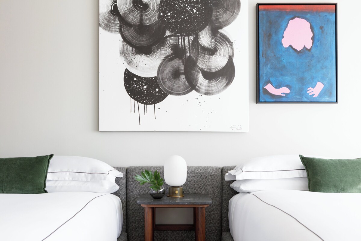 A bedroom with two beds, a small bedside table with a lamp, and a wall art piece.