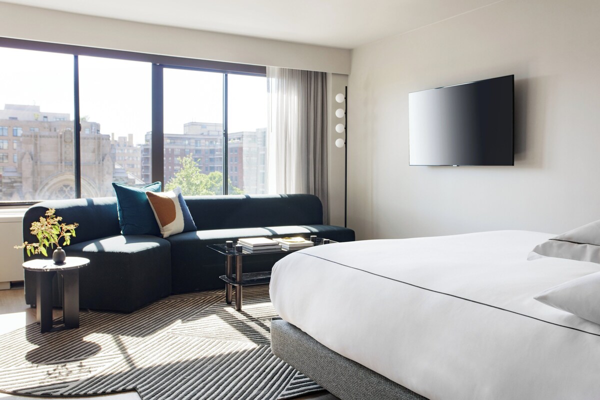 A modern hotel room with a large bed, a sofa, a coffee table, and a TV mounted on the wall.