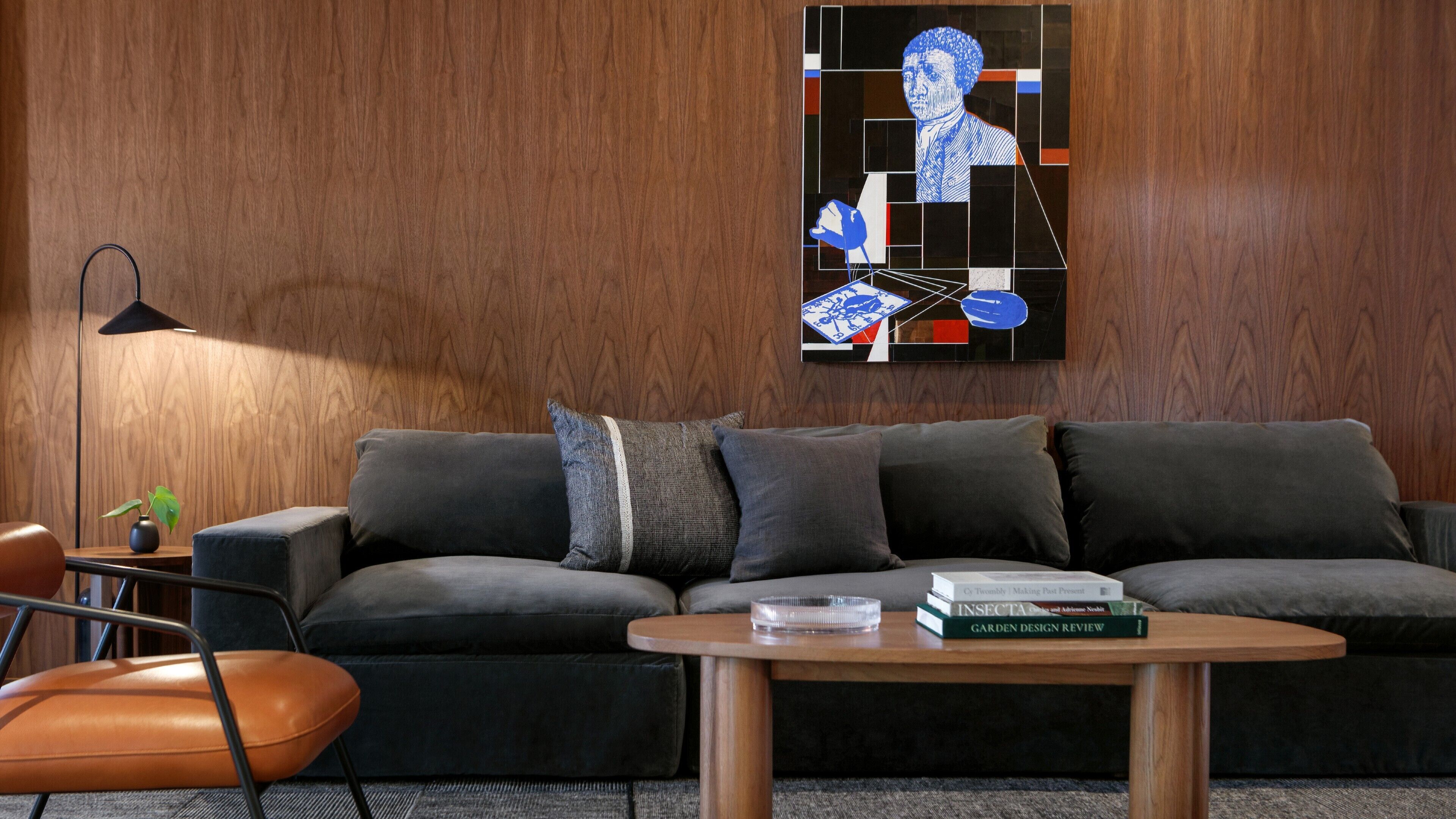 Una sala moderna con un sofá gris oscuro, varios cojines, una mesa de centro de madera con libros, una lámpara de pie y una obra de arte en la pared.