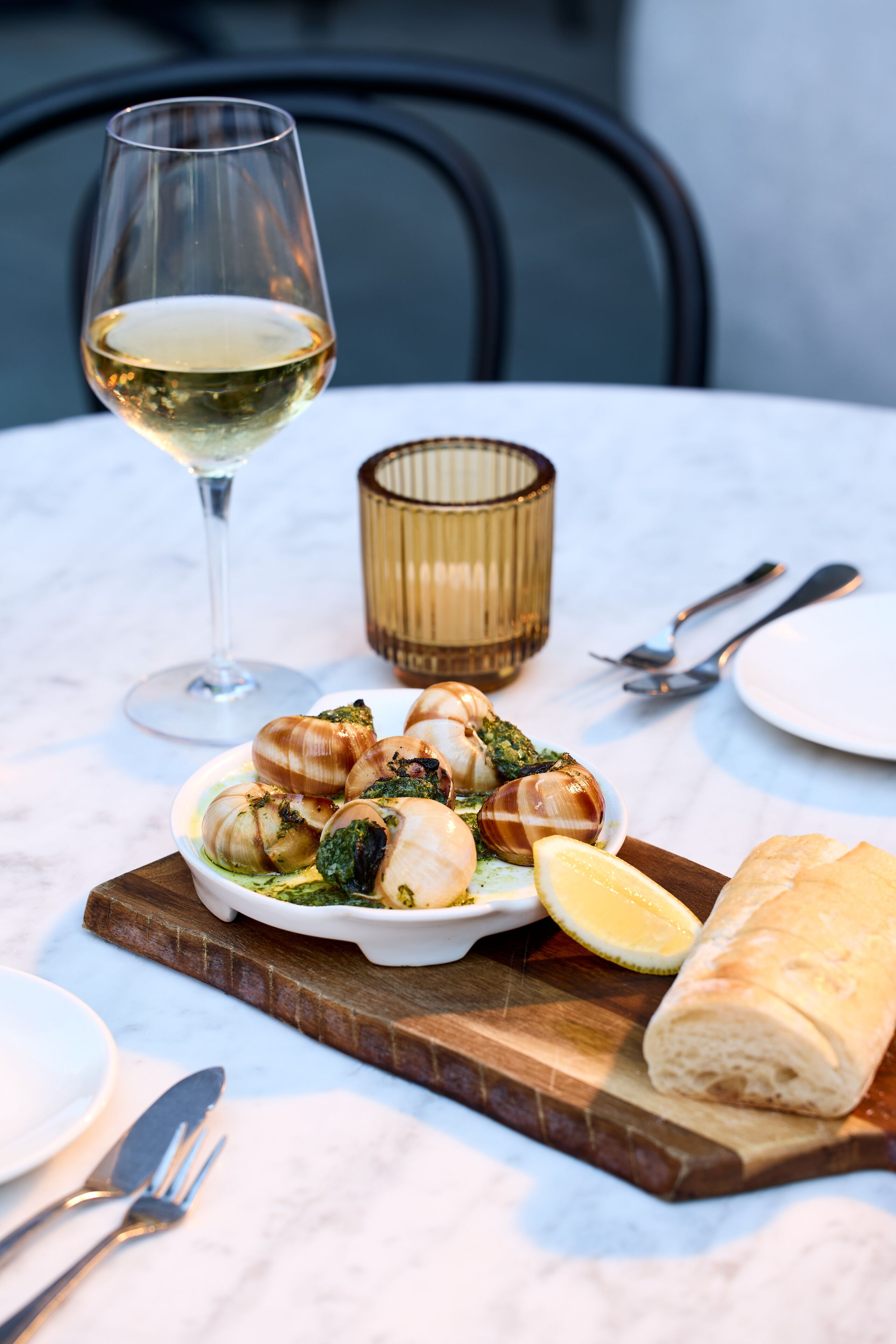 A dining table with a plate of escargot, a glass of white wine, and a loaf of bread on a wooden board.