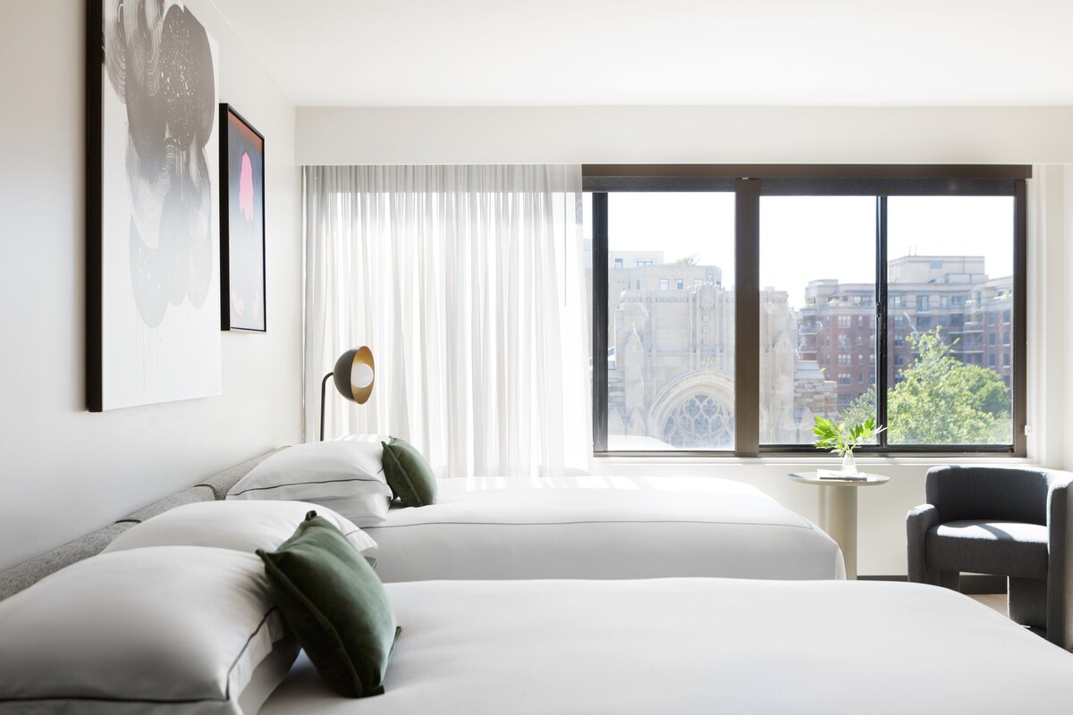 Habitación de hotel con dos camas, un escritorio y una silla. Amplia ventana con vistas a edificios y árboles.