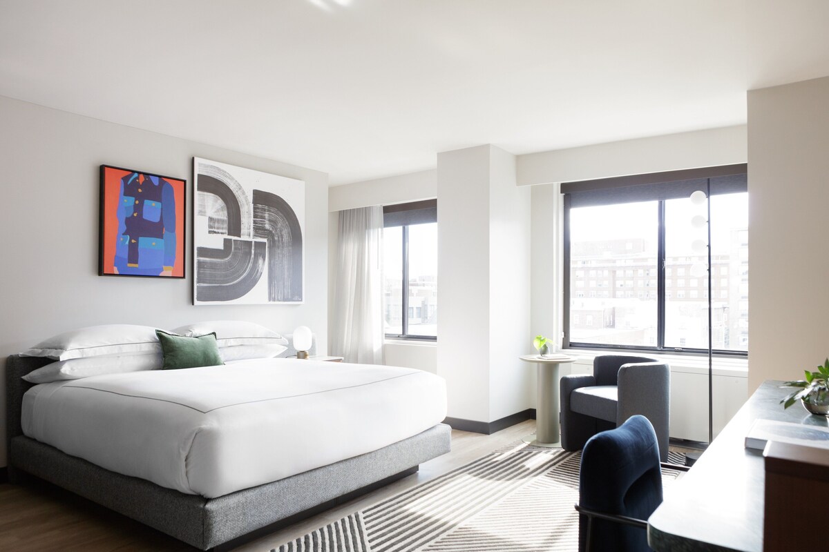 Una habitación de hotel moderna con una cama grande, un escritorio y vistas de la ciudad a través de amplios ventanales.