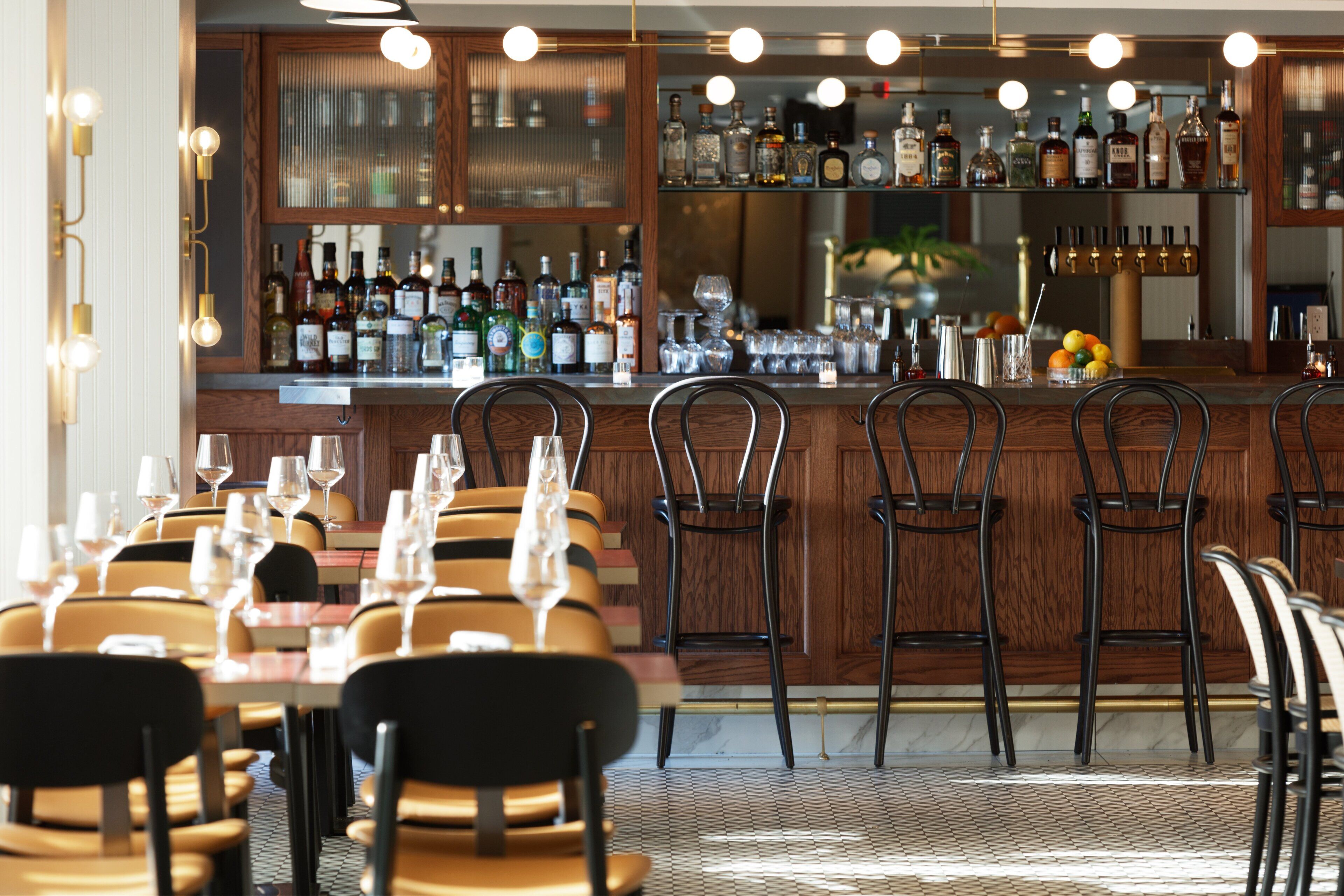 L’intérieur d’un restaurant, avec un coin bar, des chaises en bois et diverses bouteilles et verres exposés sur des étagères.