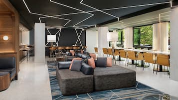 Un hall d’hôtel moderne avec un plafond au design géométrique, un canapé modulable et un coin repas avec des chaises jaunes.
