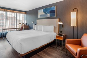 A modern hotel room with a large bed, a desk with a chair, a standing lamp, and a view of buildings through the window.