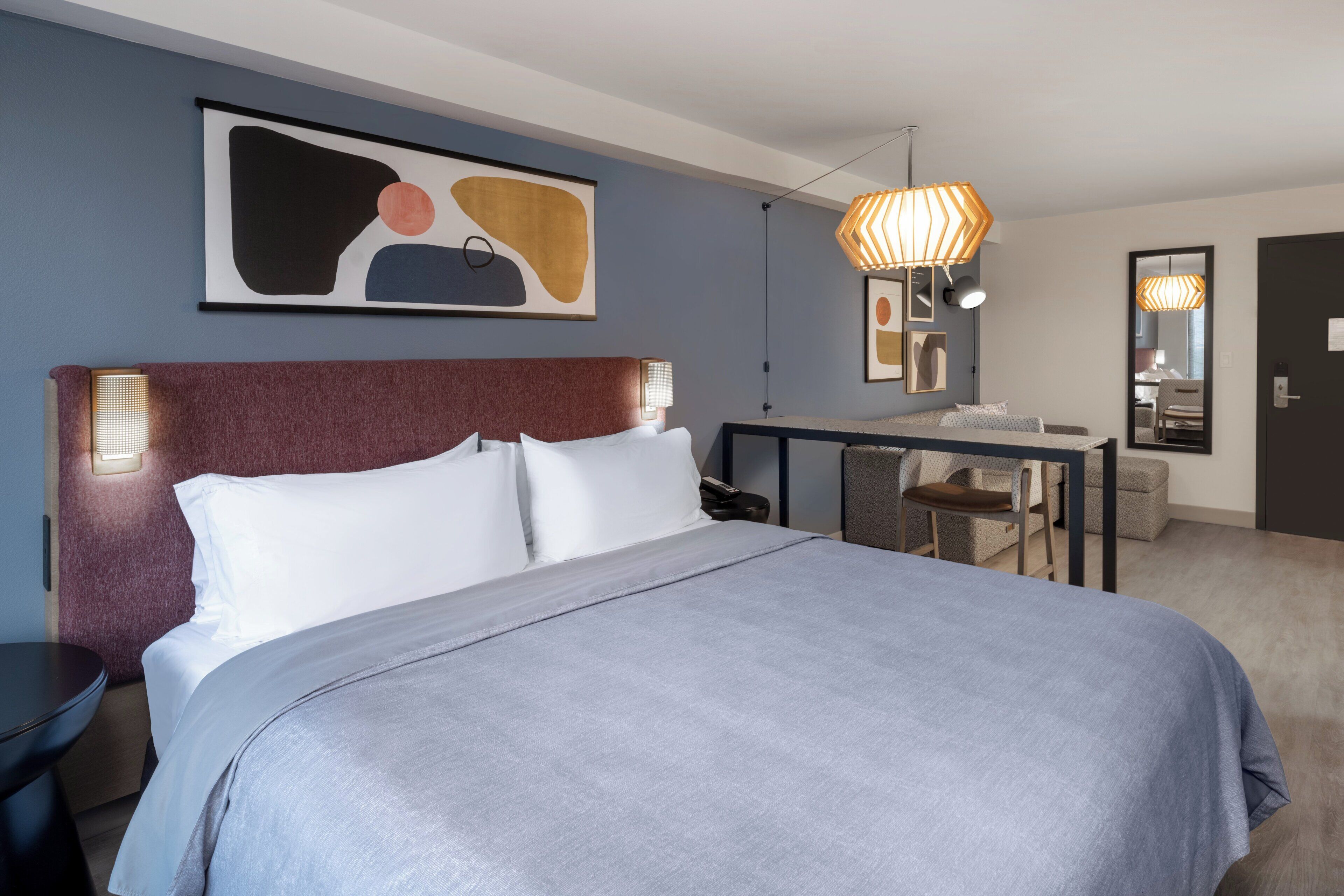 A hotel room with a large bed, bedside tables, a desk with a chair, and a hanging light fixture.