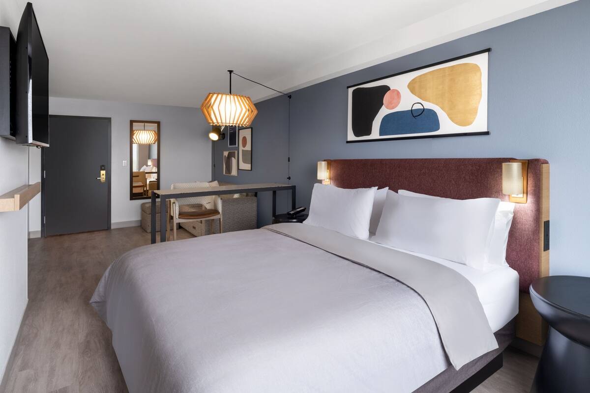 A hotel room with a bed, desk, chair, and artwork on the wall.