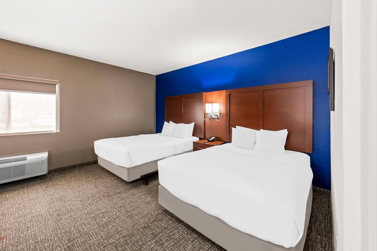 A hotel room with two beds, a large window, a wall heater, and a blue accent wall.