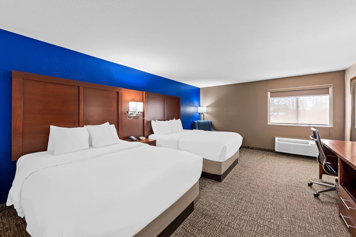A hotel room with two beds, a desk, a chair, a lamp, and a window.