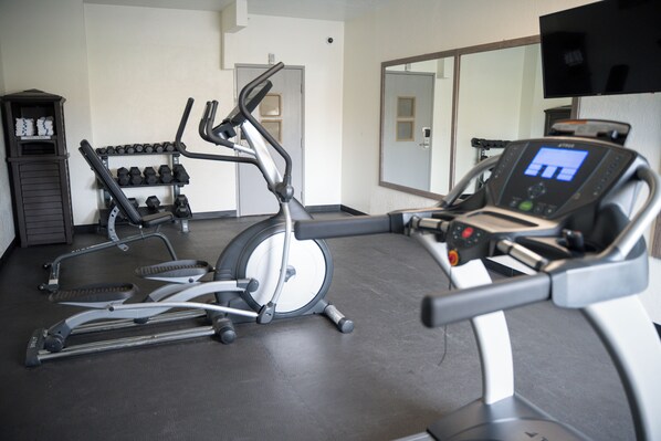 A gym with cardio machines, dumbbells, and a TV screen.
