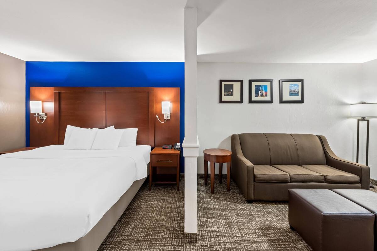 A hotel room with a bed, a brown sofa, a small table, and a nightstand. There are wall-mounted lights and framed pictures on the wall.