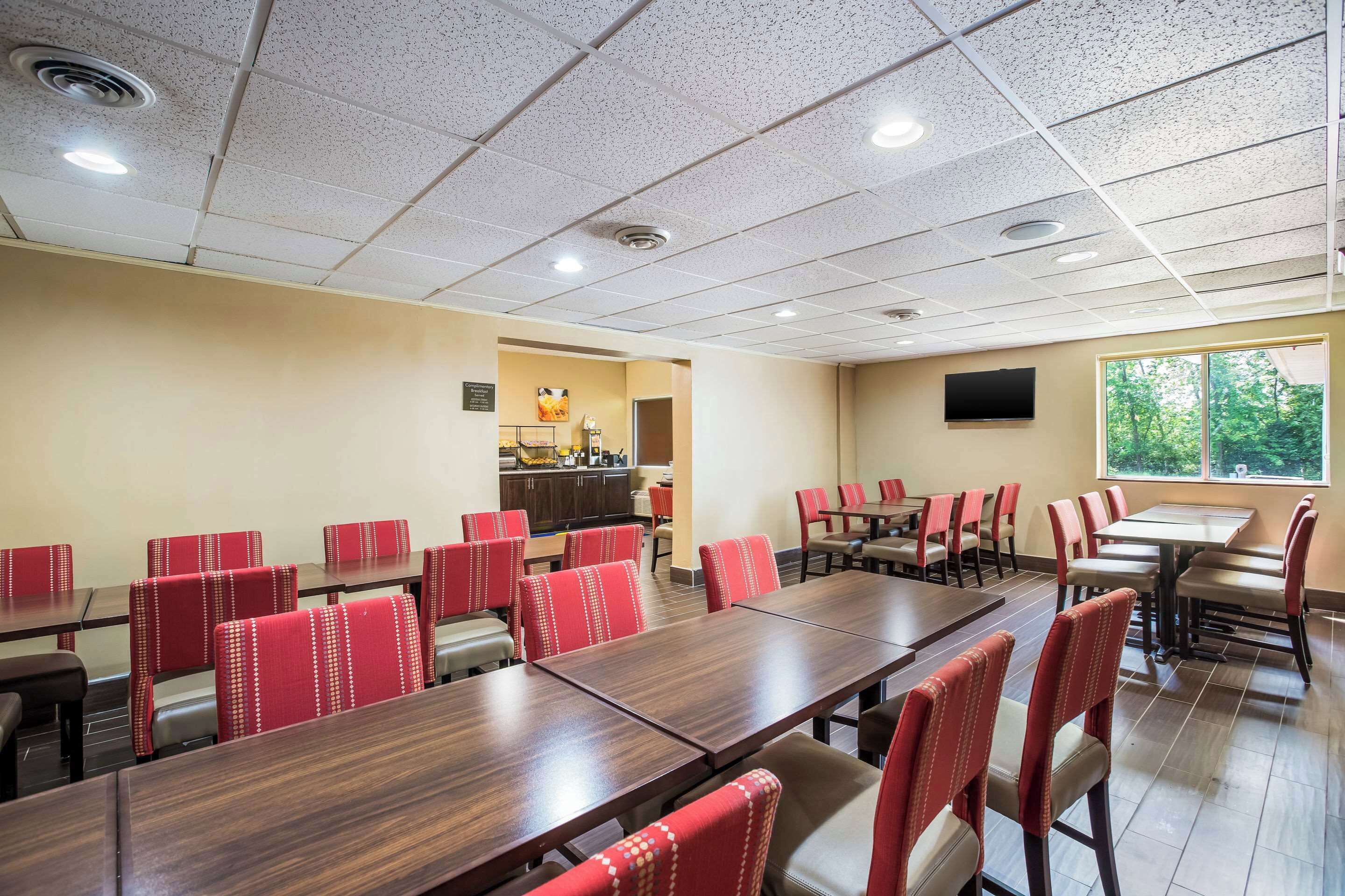 A conference room with multiple tables and chairs, a kitchenette, and a large window with a view of trees.