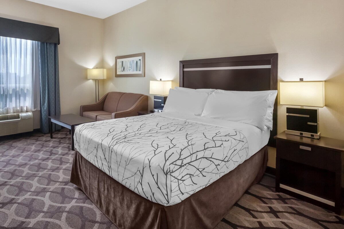 A hotel room with a large bed, a desk, a chair, a nightstand, a lamp, and a window with curtains.