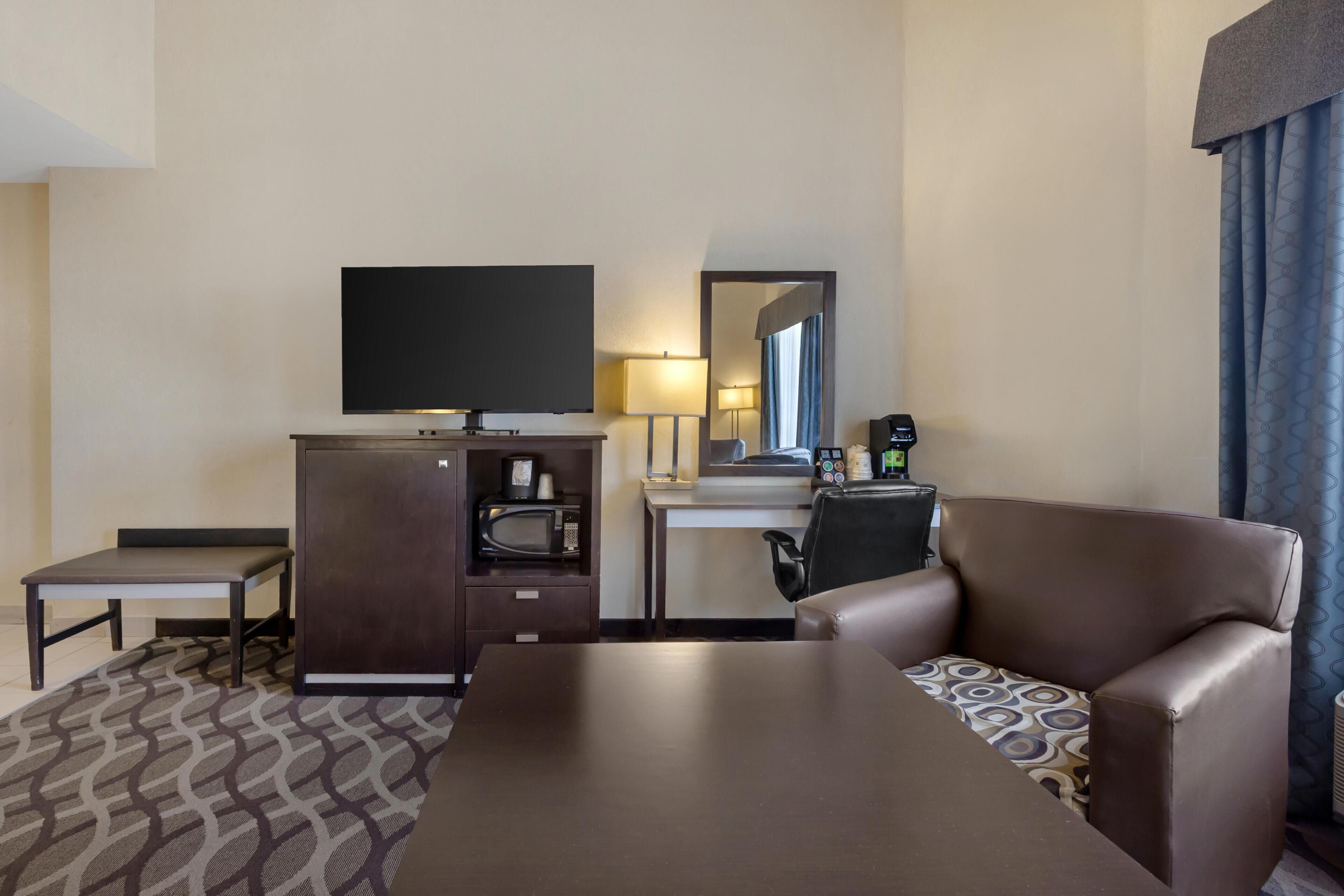 A hotel room with a flat-screen TV, a desk with a lamp, a microwave, a coffee maker, a sofa, and a patterned rug.