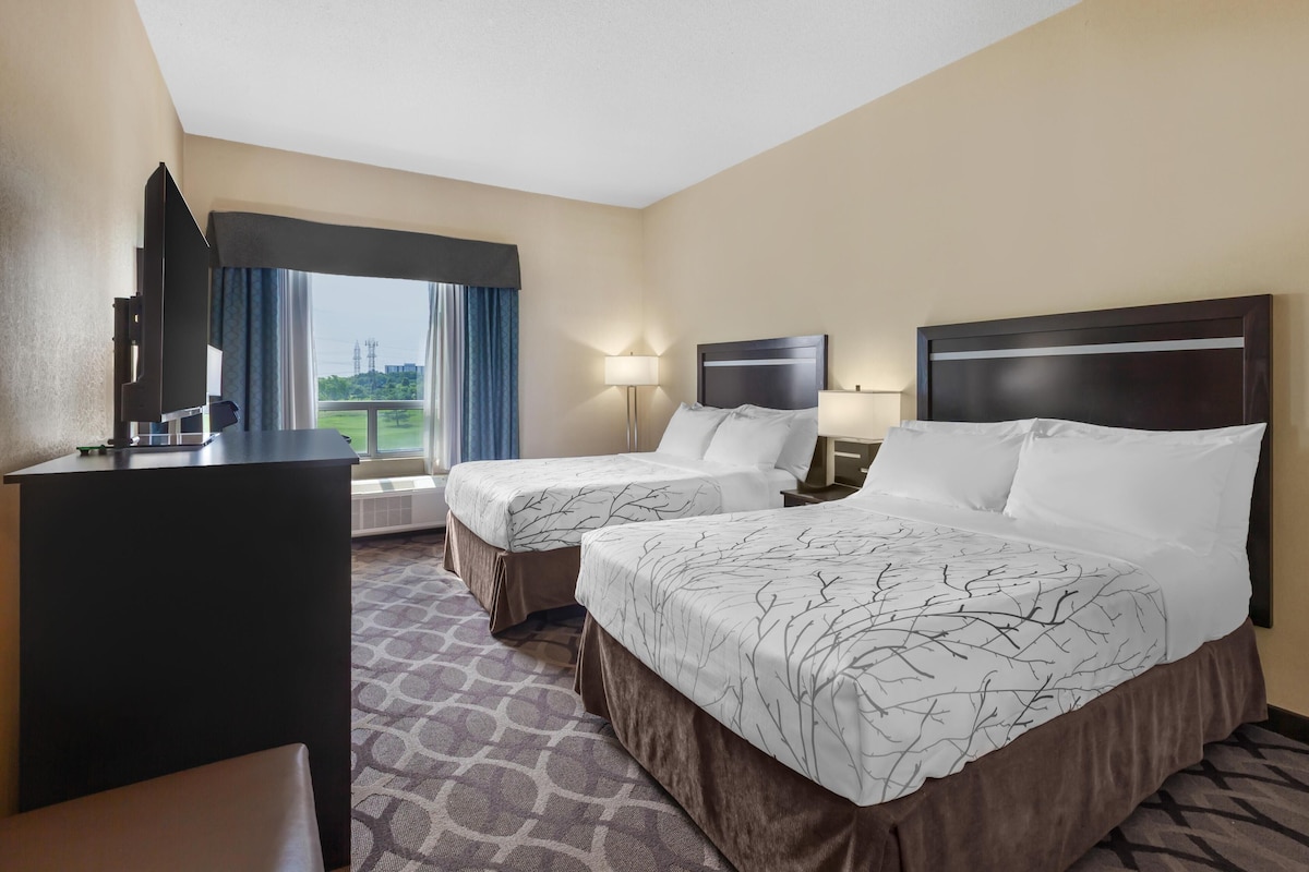 A hotel room with two beds, a desk, a chair, a window with curtains, and a view of the outdoors.