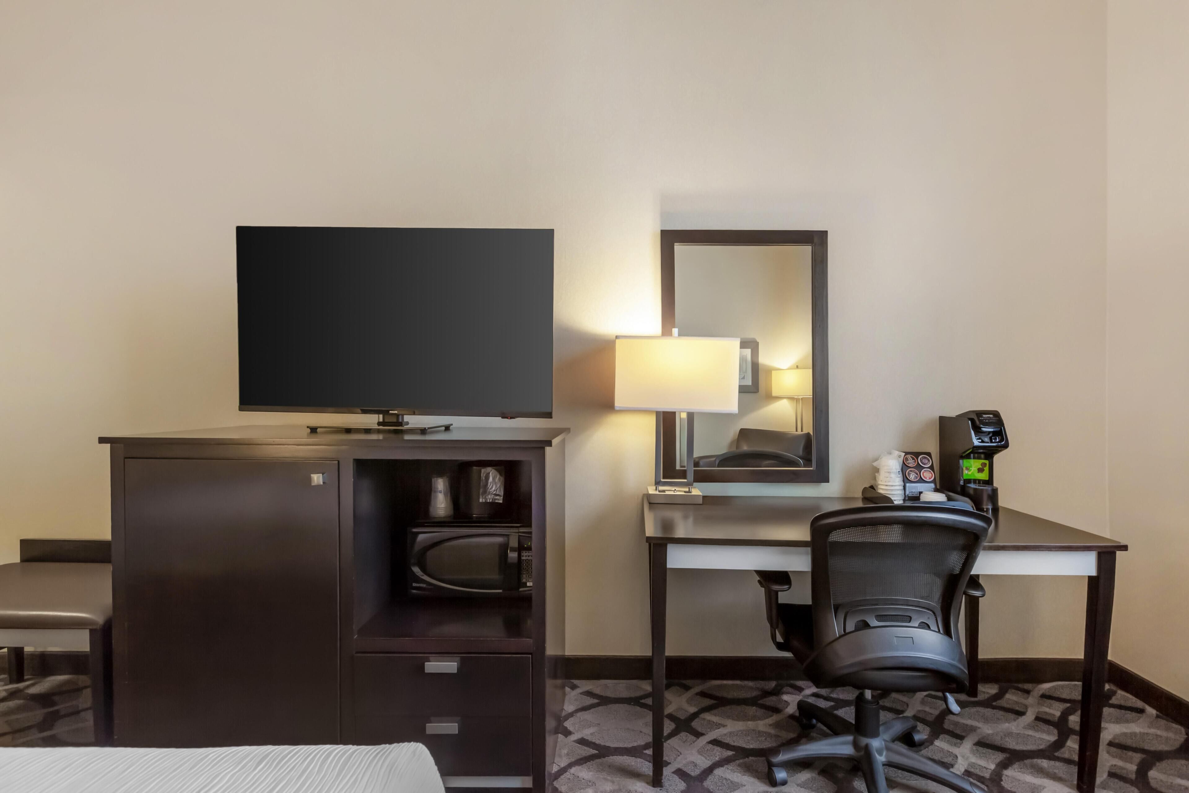 A hotel room with a bed, a desk with a chair, a television, a microwave, a coffee machine, and a mirror.